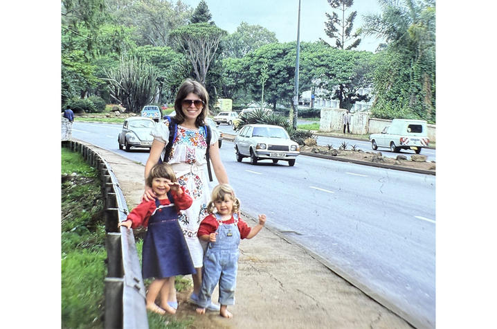 Adult standing on a sidewalk with two young children near a busy road lined with cars, trees, and greenery.