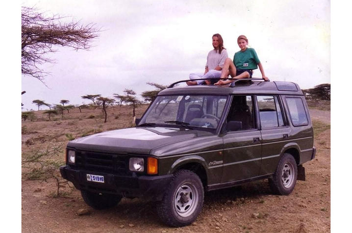 Two people sitting on the roof of a green SUV parked on a dirt road in an open landscape with scattered trees.
