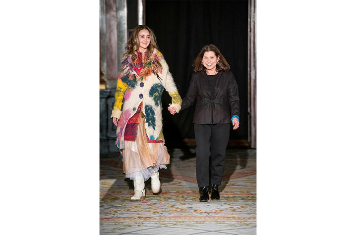 Two people walking hand in hand on a patterned runway, one wearing a colorful layered coat and boots, the other in a black outfit.