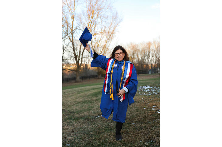 Person wearing a blue graduation gown with honor cords and a red, white, and blue stole, holding a graduation cap in one hand while standing on a grassy area with bare trees in the background.
