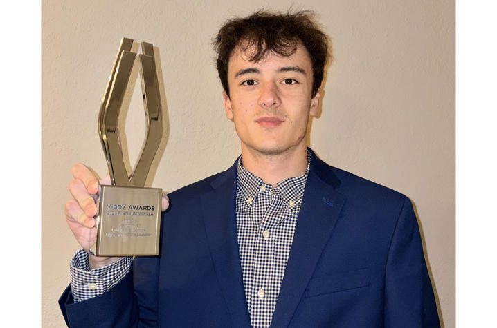Alex Masterson wearing a navy blazer and checkered button-down shirt stands against a neutral wall, holding up a metallic Viddy Awards trophy and looking at the camera with a slight smile.