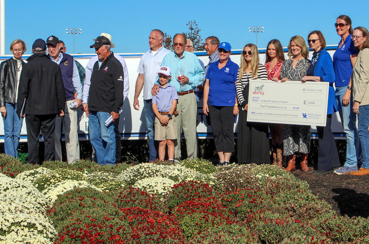 A group poses outdoors behind flower beds while several people hold an oversized $5,000 check from Red Mile for the UK MGCAE Ag Equine Programs’ Lexington Trot Breeders Association Student Scholarship.