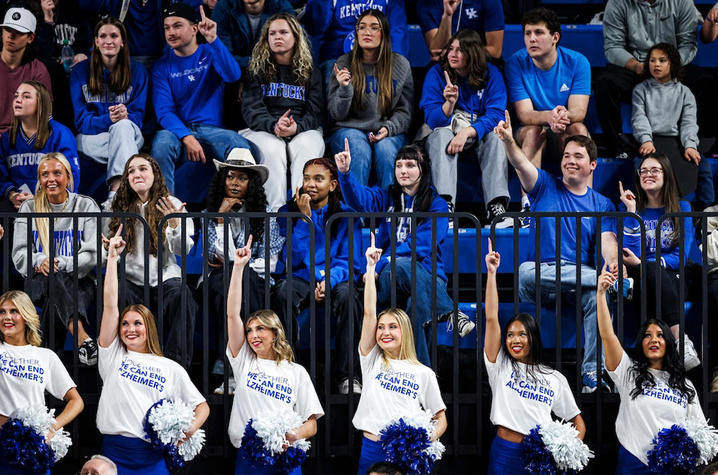 Cheerleaders wearing white “Together We Can End Alzheimer’s” shirts perform on the sidelines, holding blue and white pom-poms and raising one finger in the air. Behind them, fans dressed in blue fill the arena stands during a game.