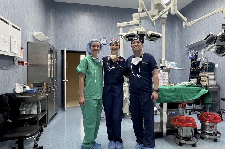 Surgical team members in an operating room during a medical mission trip Three members of a surgical team wearing scrubs, surgical caps and shoe covers stand together in an operating room surrounded by surgical lights, medical equipment and a covered operating table.