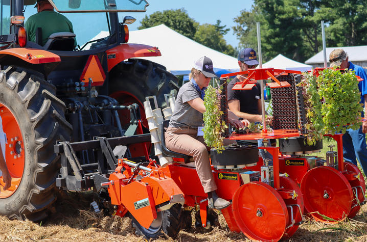 Checchi & Magli’s Trium no-till transplanter A large orange tractor is attached to a red vegetable transplanter during an outdoor agricultural equipment demonstration. Several people are gathered around the machine, with one person seated at the planting station loading seedling trays.