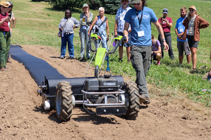 demonstration of Earth Tools’ A field day participant demonstrates a walk-behind agricultural implement that lays black plastic mulch over a raised planting bed. The machine moves down a freshly prepared row while a group of attendees stands along the edge of the field