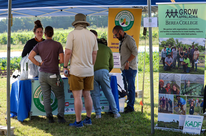 Field day attendees Several attendees gather beneath a blue canopy tent at an agricultural field day information booth. Participants stand around a table speaking with exhibitors and viewing educational materials. Large banners present.