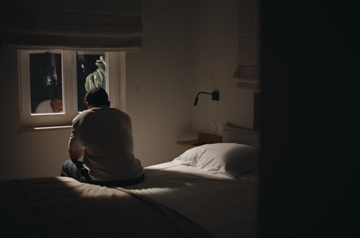 A person sits on the edge of a bed in a dimly lit bedroom, facing a small window with curtains partially open. Light from outside outlines the figure, while the rest of the room—bed, pillow, and wall-mounted lamp—remains mostly in shadow.