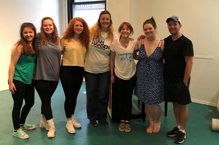 A group of seven people stands side by side in a bright studio space with teal flooring, posing together near a large window.