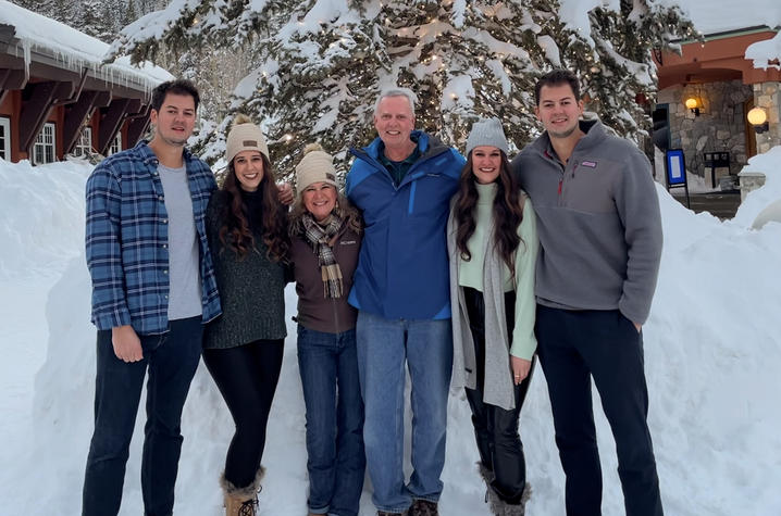Six people standing together outdoors in the snow in front of a snow-covered tree