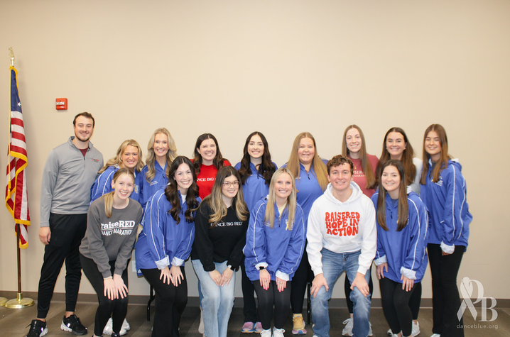 Group of young adults pose indoors against a beige wall, smiling at the camera. Most wear blue or gray casual clothing, with one man seated in front wearing a white sweatshirt. An American flag stands to the left of the group. 