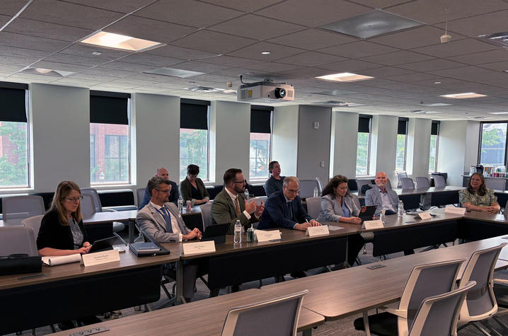 photo of KNEDA Meeting participants sit around a U-shaped conference table during a formal discussion. Several people take notes or speak while others listen in a bright room with large windows and presentation equipment.