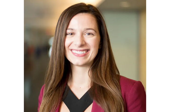 A smiling woman with long brown hair wears a burgundy blazer over a black top in an indoor portrait.