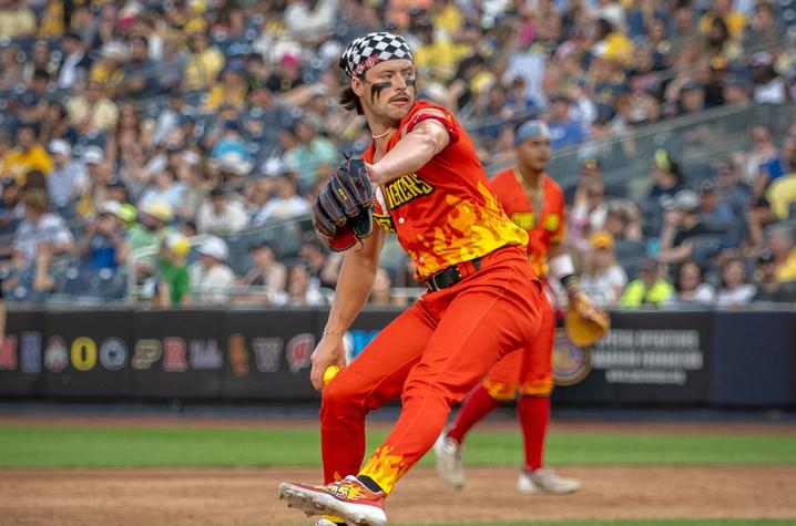 A baseball pitcher in a bright orange and red uniform throws a pitch on the mound, with a full stadium crowd blurred in the background.