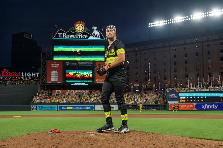 Baseball pitcher stands on the mound during a night game with a packed stadium and scoreboard lit behind him.