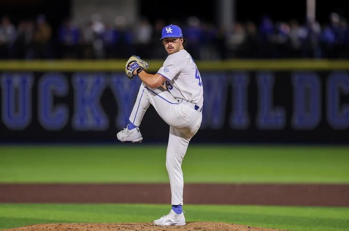 A University of Kentucky baseball pitcher in a white Wildcats uniform delivers a pitch on the mound during a night game.