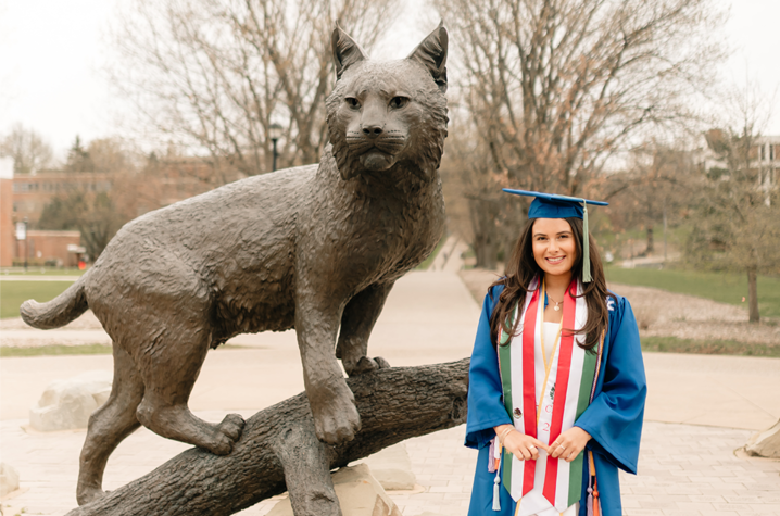 Monce standing in her graduation regalia next to one of the Wildcat statues, Bowman.