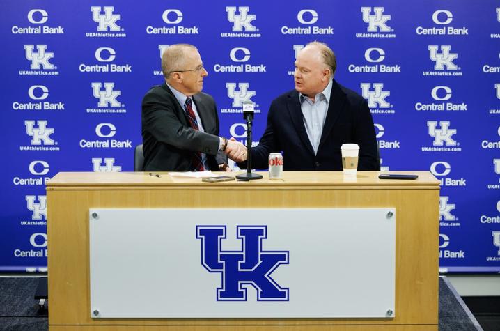 image of Dr. Scottie Day and Coach Mark Stoops shaking hands in front of a blue UK branded background