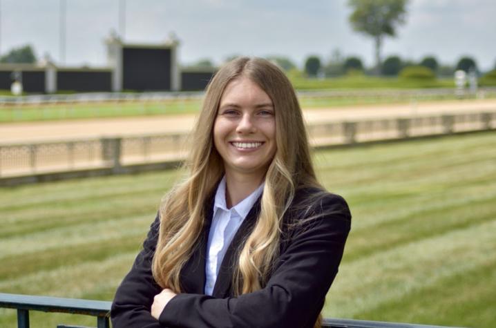 photo of Carly Schuerger A person with long hair stands outdoors with arms crossed, wearing a dark blazer over a white shirt, with a grassy field and railing in the background.