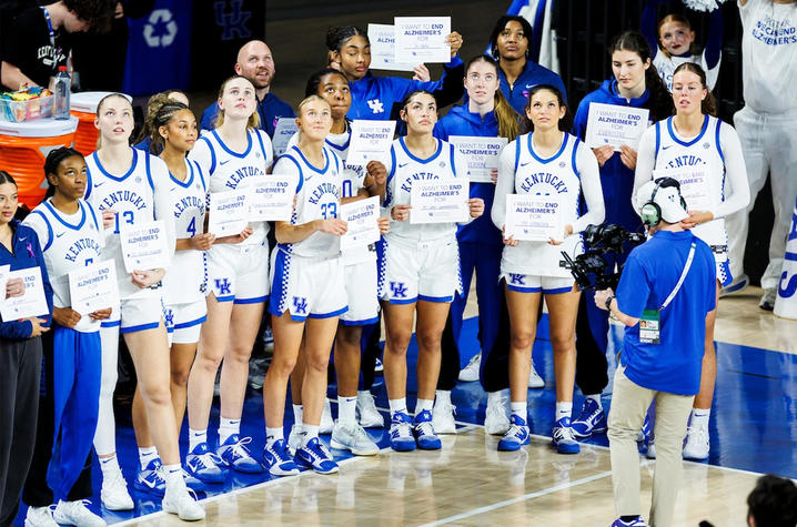 Members of the University of Kentucky women’s basketball team stand together on the court holding signs that read “I want to end Alzheimer’s for …” during a game-day event. A camera operator films them as they look upward, with teammates and staff.