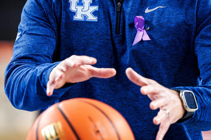 A close-up shows a person in a blue University of Kentucky pullover with a purple awareness ribbon pinned to the chest, reaching toward a basketball midair. The focus is on the hands and ball, with a gym setting softly blurred in the background.
