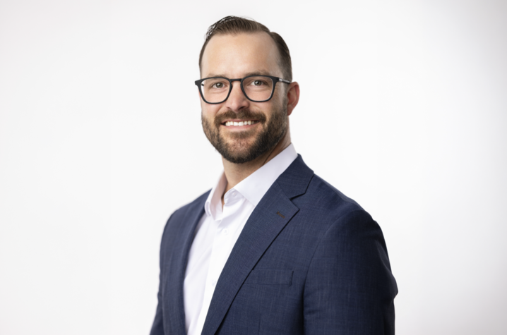 photo of Sean Schaub Studio portrait of a smiling adult with glasses and a trimmed beard, wearing a blue suit jacket and white dress shirt. The person faces the camera against a light gray background.