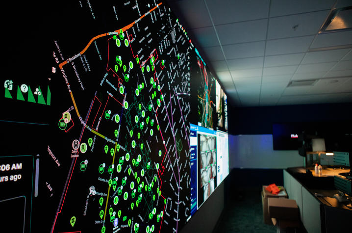 A darkened control room with a wall-sized digital map displaying green data points and lines, surrounded by monitors and workstations.