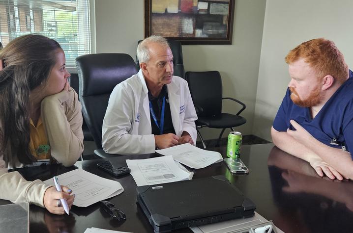 Doctor in white coat at center sitting at a table with a female resident on his left and male resident on right.