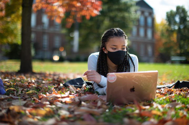 female student wearing mask lying in fallen leaves looking at her computer in front of the Main Building.