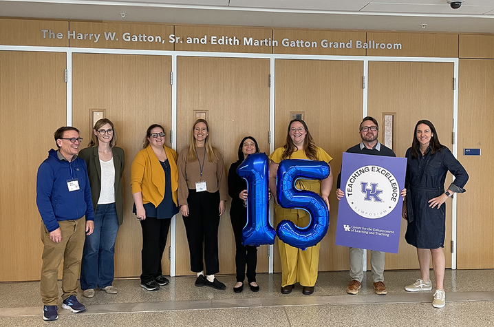 A group of eight people stand in a row in front of wooden double doors labeled “The Harry W. Gatton, Sr. and Edith Martin Gatton Grand Ballroom.” Two people in the center hold large blue balloons shaped like the number 15.