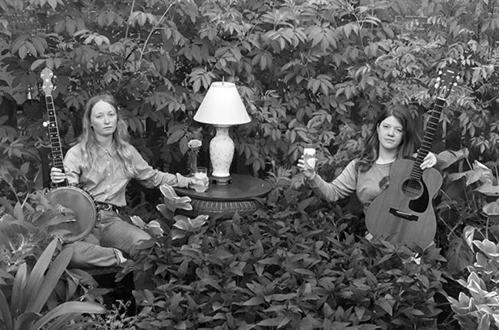 black and white photo of the duo The Other Years seated with banjo and guitar by side table with lamp in overgrown field