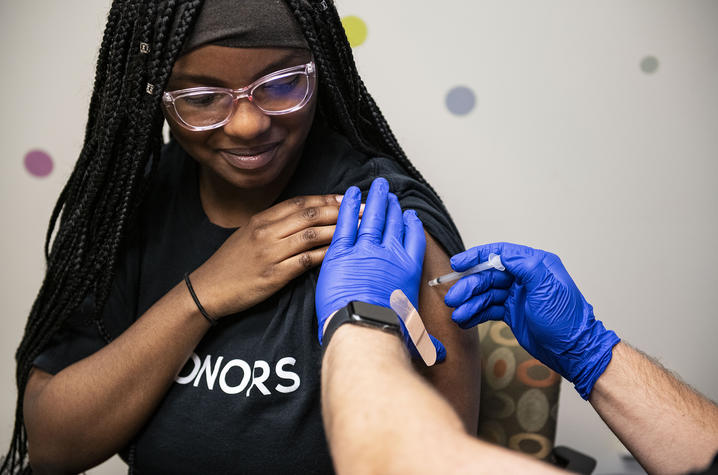 A health care worker wearing blue gloves administers a vaccine injection into a person's upper arm while the person holds up a shirt sleeve.