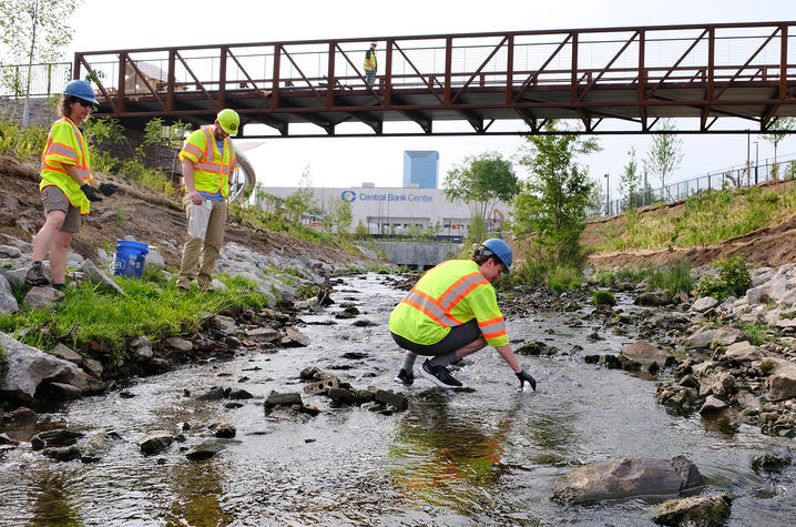 Three researchers in high-visibility safety vests and hard hats collect water samples in a shallow rocky stream beneath a metal pedestrian bridge, with stabilized creek banks and a commercial building visible in the background.