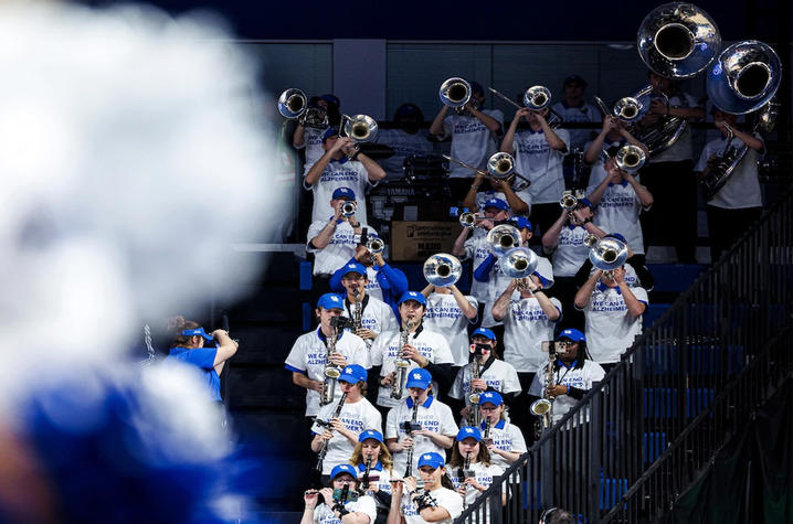 Members of a pep band wearing blue caps and white “Together We Can End Alzheimer’s” shirts play brass and woodwind instruments in arena seating. The musicians perform from the stands, with instruments raised and a blurred figure in the foreground.