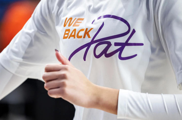 A close-up shows a person wearing a white shirt that reads “We Back Pat” in orange and purple lettering. The person gives a thumbs-up gesture, with the focus centered on the message across the chest.