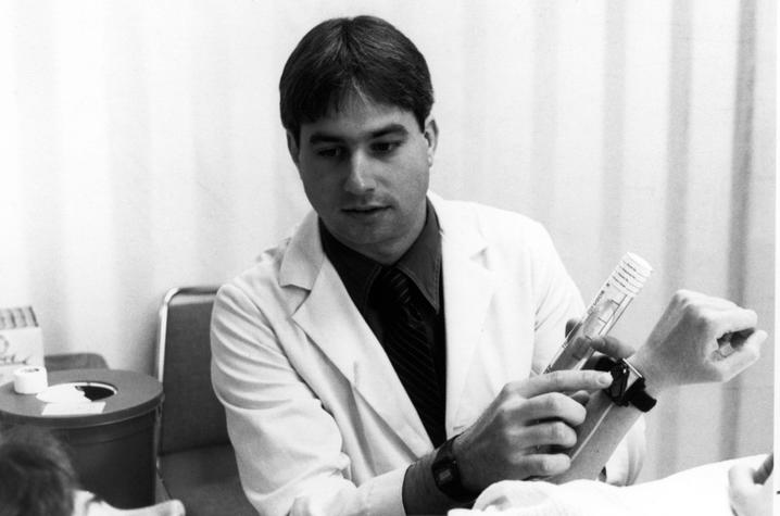 A provider in a white lab coat demonstrates how to use an injection device on a patient’s forearm in a clinical setting. Medical equipment and a treatment chair are visible nearby, emphasizing a hands-on teaching or research moment.