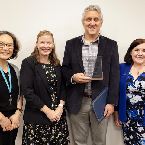Four people in a group photo, with one person holding a glass award and a blue certificate folder against a plain indoor background.