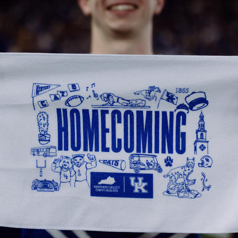 Close-up of a person holding a white rally towel featuring a blue “Homecoming” design with University of Kentucky branding and illustrated campus-themed icons. A blurred crowd and another person are visible in the background.