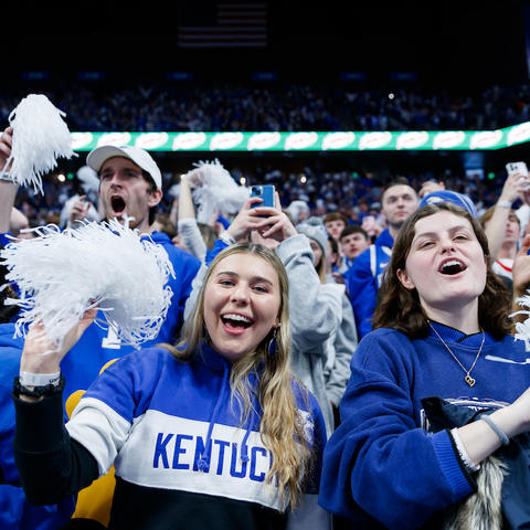 A group of spectators in blue Kentucky shirts stand in an indoor arena, raising white pom‑poms and cheering. The crowd is tightly packed, with bright arena lighting and a scoreboard glow visible in the background.