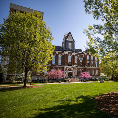 The front of Main Building at the University of Kentucky during the springtime, with trees and grass in the foreground and a clear blue sky in the background.