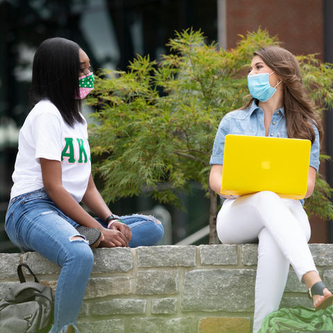two female students wearing masks sitting on wall outside talking to each other