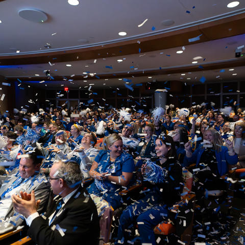 A large group of people seated in an auditorium, wearing blue and white attire and accessories such as scarves and pom-poms. Blue and white confetti fills the air, creating a festive atmosphere. 