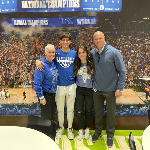 Four people pose together in front of a large mural depicting a University of Kentucky basketball championship celebration. The group stands with their arms around one another, wearing Kentucky-themed clothing, confetti and a packed arena crowd in backgro