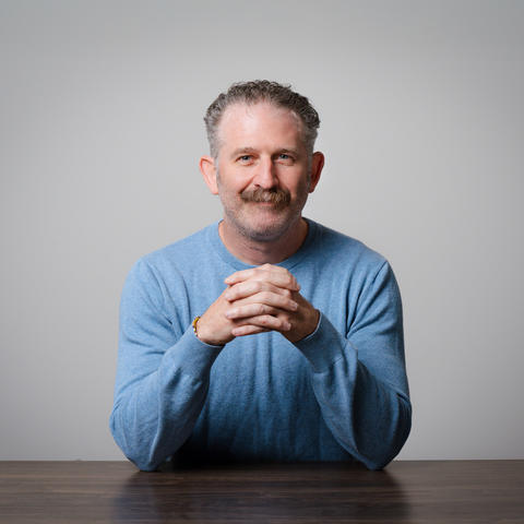 Studio portrait of an adult with short curly hair and a mustache, seated at a table with hands clasped. The person wears a light blue sweater against a plain light gray background.