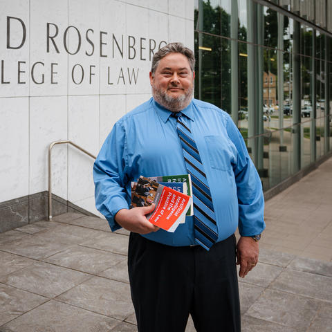 A person stands outside the Rosenberg College of Law building, holding several books.