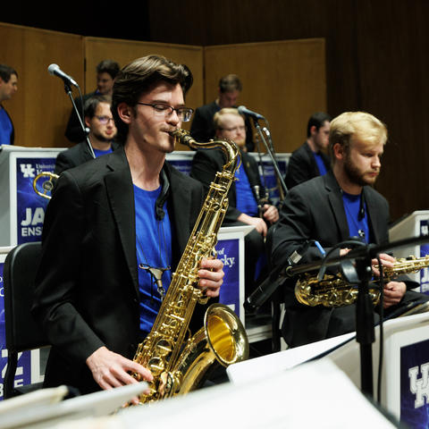 white male in a black jacket and blue shirt playing tenor saxophone in a jazz ensemble
