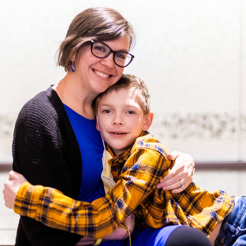 Image of Dynae Utz in blue dress with black sweater, hugging her nine-year-old son Lewis who is wearing a yellow plaid shirt and jeans.