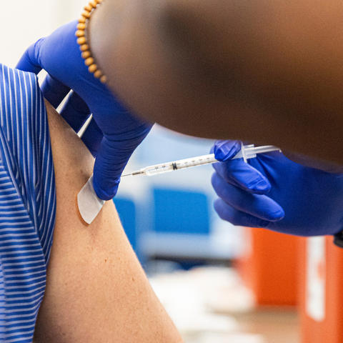A health care worker wearing blue gloves gives a vaccine injection in the upper arm of a person wearing a blue UK shirt. A small bandage is placed near the injection site.
