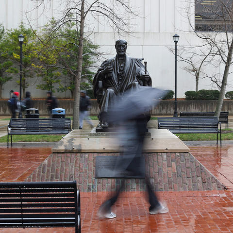 A photo of a student walking in front of the Patterson statue in the rain with a black umbrella.