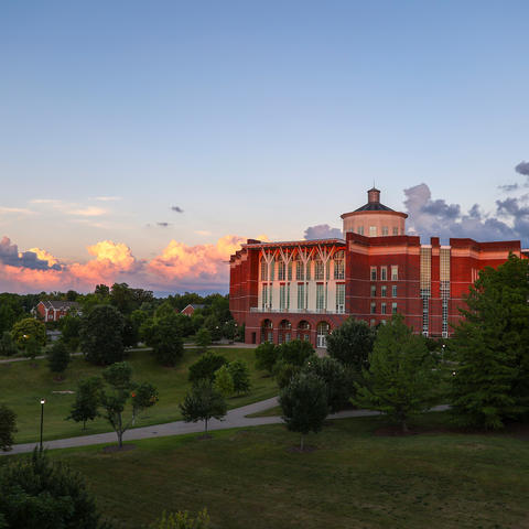 Red-brick campus building seen at sunset with trees and open green space in the foreground and colorful clouds in the sky.